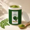 Open jar of Organic Moringa Powder showing green powder inside with fresh moringa leaves and a wooden spoon nearby.
